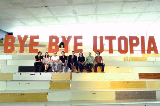 This picture shows eight members of Raumlabor Berlin sitting underneath a wooden lettering reading "BYE BYE UTOPIA".